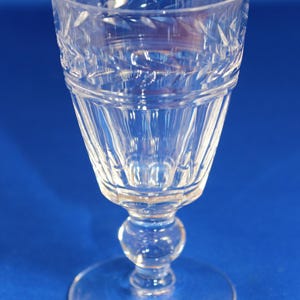 May include: A clear crystal wine glass with a round base and a decorative stem. The bowl of the glass has an etched pattern and vertical lines. The glass is transparent and stands out against a blue backdrop.
