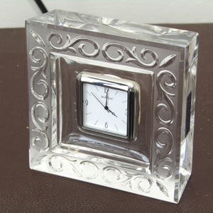 Puede incluir: Un reloj de cristal transparente con un diseño ornamentado y una esfera plateada. La esfera del reloj tiene marcas de números romanos negros y agujas negras.
