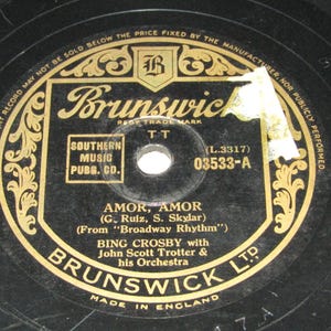 May include: A black vinyl record with gold and white text. The record label features the Brunswick logo and the song title "AMOR, AMOR" by Bing Crosby. The label also includes the text "MADE IN ENGLAND".