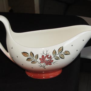 May include: White ceramic gravy boat with a red and green floral design. The boat has a wide, rounded base and a handle on the left side.
