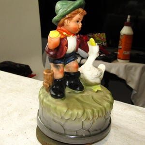 May include: A vintage porcelain music box featuring a boy in a green hat and brown jacket holding a yellow ball, standing next to a white goose. The music box has a green base with a brown wooden fence and a gray stone base.