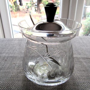 Könnte beinhalten: Eine klare Zuckerdose aus Glas mit einem silbernen Deckel und einem schwarzen Griff. Die Schüssel hat ein geätztes Blumendesign. Ein kleiner Silberlöffel befindet sich im Inneren. Die Zuckerdose ist wahrscheinlich Vintage.