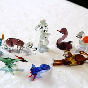 May include: A collection of ten glass animal figurines, including a blue rabbit, a purple dog, a clear poodle, a purple swan, a clear bird with orange accents, a brown cat, a brown dog, a green bird, and a blue bird.