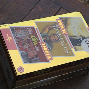 Op de afbeelding: Een drievoudig pakket VHS-films met "El Cid", "Spartacus" en "Gladiator". De doos is geel met gouden accenten en toont afbeeldingen van de acteurs uit elke film. Het prijskaartje geeft "£11.99" aan.