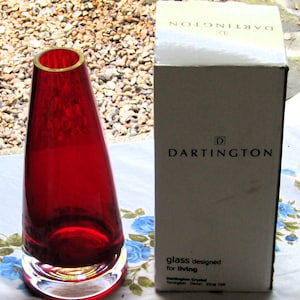 May include: A red glass vase with a gold rim and a clear glass base. The vase is in its original box, which is white with black text that reads "Dartington glass designed for living".