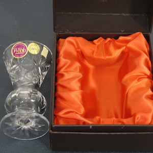 May include: A clear crystal Webb vase with a starburst base and diamond-cut pattern, next to a brown box lined with orange satin. The vase has two stickers on it. The box is open, revealing the satin lining.