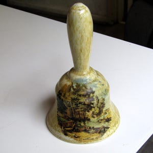 Puede incluir: Una campana de cerámica con un asa beige y una escena pintada de un paisaje campestre. La campana tiene un esmalte marrón claro con un borde blanco.