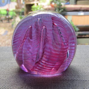 Puede incluir: Un pisapapeles de cristal transparente con un diseño rosa y morado vibrante. El interior presenta patrones arremolinados y líneas verticales, creando un efecto visual único. La forma esférica se asienta sobre una superficie texturizada.