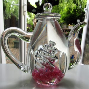 May include: A clear glass teapot with a spiral design inside and a pink base. The teapot has a handle, a spout, and a lid with a small knob. The background is a blurred view of a garden.