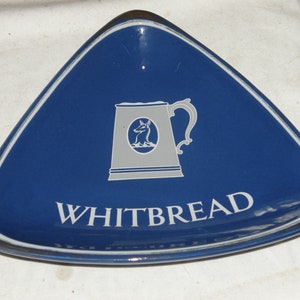 May include: A blue triangular ceramic ashtray with a white outline. The ashtray features a white image of a beer mug with a stag inside and the text "WHITBREAD" in white.