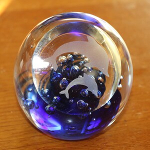 Puede incluir: Un pisapapeles de cristal transparente con un interior azul y un diseño de delfines grabado. El pisapapeles tiene forma esférica y contiene burbujas de aire. Los delfines están grabados en color blanco esmerilado.