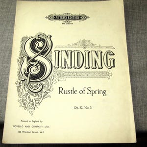 以下が含まれることがあります： 「Rustle of Spring」と書かれた楽譜。装飾的な文字で「SINDING」と書かれています。表紙はクリーム色で、黒い文字と装飾的な要素があります。イギリスでNovello and Company, Ltd.によって印刷されました。