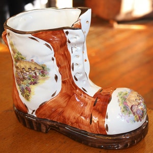 May include: A ceramic boot-shaped vase with a brown and white painted design. The vase features a cottage scene with a thatched roof and flowers.