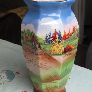 May include: A vintage ceramic vase with a hexagonal shape and a blue sky and landscape scene. The vase features a brown base, a green and yellow landscape, and a blue sky with white clouds. The scene depicts figures in period clothing.
