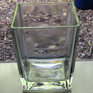 May include: A clear, square glass vase with a slightly flared shape. The vase has a thick base and a smooth, transparent surface. The edges are rounded, and the vase is empty. The background is blurred, with hints of green and brown.