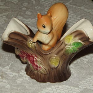 May include: A ceramic double vase shaped like a tree trunk with a squirrel figurine perched on top. The vase is decorated with brown, green, and yellow paint, and the squirrel is painted in shades of brown and white.
