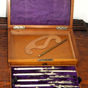 May include: A wooden box with a purple velvet lining, containing a set of vintage drafting tools. The tools include compasses, rulers, and a protractor. The box is open and the tools are arranged neatly inside.