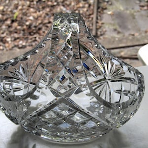 May include: A clear cut glass basket featuring a prominent arched handle and a decorative pattern of intersecting diamond shapes and radiating starburst motifs. The glass surface is highly reflective with sharp, geometric facets throughout.
