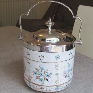 May include: A vintage ice bucket with a silver lid and handle. The white ceramic body is decorated with blue, red, and gold floral patterns. The handle is arched and attached to the sides of the bucket.
