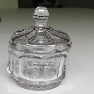 Peut inclure: Un pot en verre clair et facetté avec un couvercle. Le pot a une forme octogonale avec des panneaux décoratifs et un couvercle détaillé. Le couvercle a un dessus arrondi. Le pot est probablement destiné à ranger de petits objets ou comme pièce décorative.