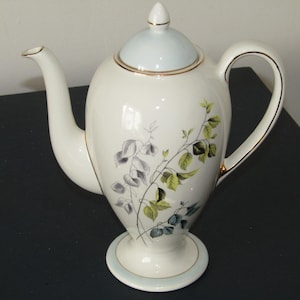 Può includere: Una teiera in ceramica bianca con coperchio e base azzurri, ornata da bordi dorati. La teiera presenta un beccuccio e un manico curvi ed è decorata con un motivo floreale nei toni del grigio, verde e blu.