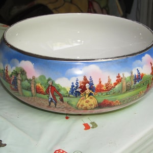May include: A round, white ceramic bowl with a painted landscape scene around the exterior. The scene depicts figures in period clothing in a garden setting with trees and a blue sky. The rim is trimmed with silver.