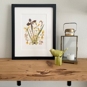 May include: A framed pressed flower art print with a black frame and a white mat. The print features a purple iris with yellow and pink wildflowers. The print is sitting on a wooden shelf with a small green bird figurine and a glass lantern.