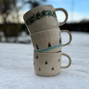 Könnte beinhalten: Drei Keramikbecher, die übereinander gestapelt sind. Der obere Becher hat ein grünes und goldenes Blattdesign. Der mittlere Becher hat ein blaues und weißes Design mit kleinen Bäumen. Der untere Becher hat einen weißen Hintergrund mit kleinen grünen Bäumen.