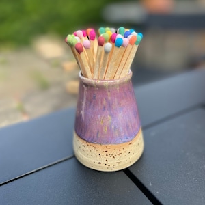 Könnte beinhalten: Ein Keramikkästchen für Streichhölzer mit einer violetten und braunen Glasur. Das Kästchen ist gefüllt mit Streichhölzern aus Holz mit bunten, bemalten Köpfen.
