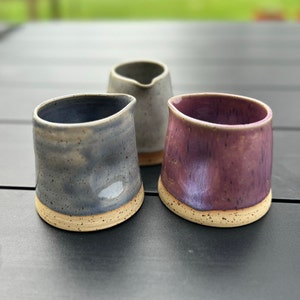 May include: Three ceramic creamer jugs in different colours. The jugs are grey, white and purple. They have a speckled glaze and a light brown rim.