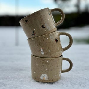 Könnte beinhalten: Drei Keramikbecher, die übereinander gestapelt sind. Die Becher sind beige mit einer gesprenkelten Glasur und mit handgemalten winterlichen Motiven verziert, darunter Schneemänner, Vögel und Schneeflocken.