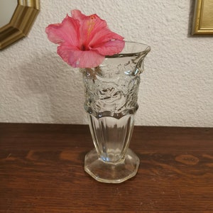 Peut inclure: Un vase en verre transparent avec une fleur d'hibiscus rose. Le vase présente un motif floral décoratif et une base hexagonale. La fleur est d'un rose vif avec des étamines jaunes.