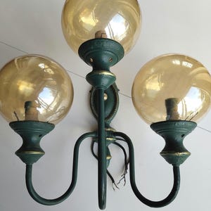 Peut inclure: Une applique murale composée de trois abat-jours sphériques en verre teinté ambre. La structure est en métal vert foncé avec des accents peints en doré sur les bords des douilles et sur la plaque de fixation centrale.