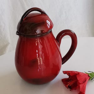 Peut inclure: Une cruche en céramique rouge brillante avec un couvercle et une anse assortis. La cruche a une forme bulbeuse et un bord marron foncé sur le couvercle. Une fleur d'hibiscus rouge repose à côté de la cruche.