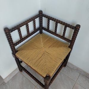 Peut inclure: Une chaise d'angle dotée d'une structure en bois brun foncé avec un design à fuseaux tournés. L'assise est constituée de paille tressée de couleur beige clair, formant un motif carré qui se rejoint au centre de la surface de l'assise.