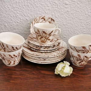 Peut inclure: Un service à thé vintage avec des tasses, soucoupes et assiettes blanches, décoré d'un motif de tiges de blé marron. L'ensemble comprend plusieurs tasses, soucoupes et assiettes, disposées sur une surface en bois foncé. Une fleur blanche est placée à côté.