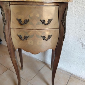 Peut inclure: Une table de chevet en bois avec un plateau en marbre, dotée de deux tiroirs avec marqueterie florale et poignées en métal ornementées. Le meuble repose sur quatre pieds galbés ornés de garnitures métalliques sur les bords avant.