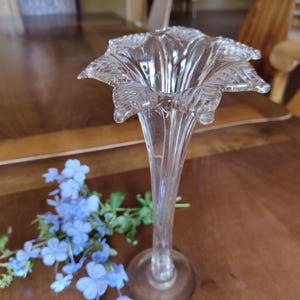 May include: A clear glass vase with a flared, flower-like top and a slender stem. The vase sits on a round base. A sprig of blue flowers rests beside the vase on a brown wooden surface. The vase is approximately 25 cm tall.