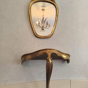 May include: Ornate gold-colored mirror with a decorative top, reflecting a chandelier. Below, a matching gold-colored wall shelf with a curved design and a single support leg. The mirror and shelf are mounted on a light blue textured wall.