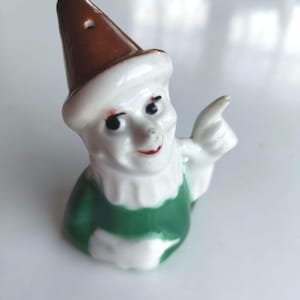 Peut inclure: Figurine de gnome en céramique avec un chapeau pointu marron, un visage blanc et une salopette verte. Le gnome a des yeux noirs, un sourire rouge et pointe avec une main. L'objet est décoratif.