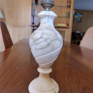 Peut inclure: Une lampe de table en marbre blanc et gris avec un design décoratif sculpté. La lampe a une douille en haut et une base ronde. Le cordon de la lampe est visible. La lampe est sur une table en bois.