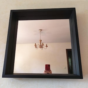 Peut inclure: Miroir carré encadré de noir. Le miroir reflète une pièce avec un lustre, une porte et une personne tenant un téléphone rouge. Le cadre est épais et de conception simple et moderne. Le mur derrière le miroir a un motif texturé.