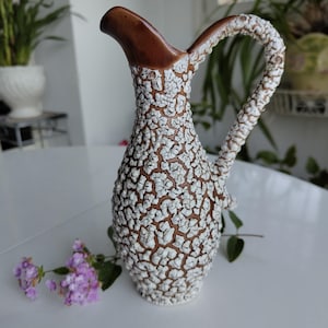 Peut inclure: Pichet en céramique marron et blanc avec une surface texturée et craquelée. Le pichet a une anse incurvée et un bec verseur. Une brindille de fleurs violettes repose près de la base. Le pichet est probablement vintage.