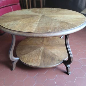 Peut inclure: Une table basse ronde en bois avec une finition brun clair et un motif de placage en éventail sur le plateau supérieur et l'étagère inférieure. La table est soutenue par quatre pieds incurvés partant du bord supérieur vers l'étagère.