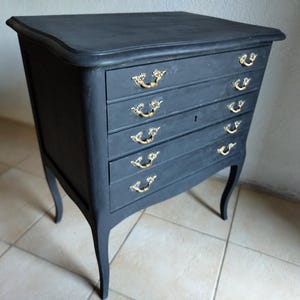 Può includere: Una cassettiera nero opaco con cinque cassetti orizzontali, ciascuno dotato di due maniglie in metallo dorato decorate. Il mobile poggia su quattro gambe curve e presenta una piccola serratura sulla parte anteriore del secondo cassetto.