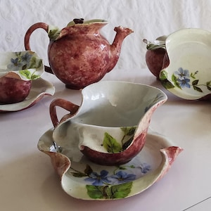 Peut inclure: Service à thé en céramique avec théière, tasses, soucoupes et pot à crème. L'ensemble présente un coloris bordeaux et blanc avec des motifs floraux. La théière a une anse en forme de feuille et un dessus décoratif.