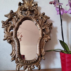 Puede incluir: Espejo de pared dorado y ornamentado con superficie ovalada. El marco presenta elaborados detalles de volutas y flores. El espejo se muestra contra una pared clara, con una maceta roja de orquídeas moradas en el fondo.