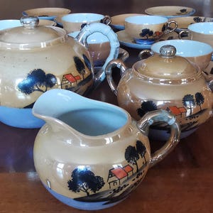 May include: A vintage tea set with a pearlescent finish, including a teapot, sugar bowl, creamer, and several teacups and saucers. The set is decorated with a landscape design of trees and houses in black and red against a gold background, with blue accents.