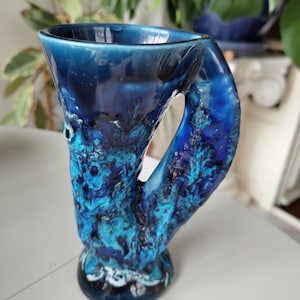 Peut inclure: Une tasse en céramique avec un bord évasé et une anse épaisse intégrée. La surface présente un vernis brillant aux tons multiples, allant du bleu marine profond au bleu roi et turquoise vif, avec une texture organique très irrégulière.