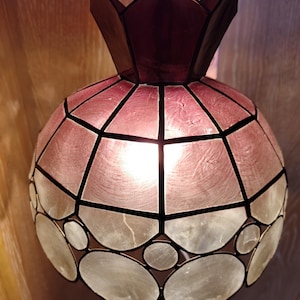 Op de afbeelding: Een decoratieve hanglamp met een uniek ontwerp. De lamp heeft een bordeauxrode bovenkant en een bolvormig lichaam met een roze-wit, glas-in-loodachtig patroon. Het ontwerp omvat geometrische vormen en cirkelvormige accenten, wat een warme, omgevingsgloed creëert.